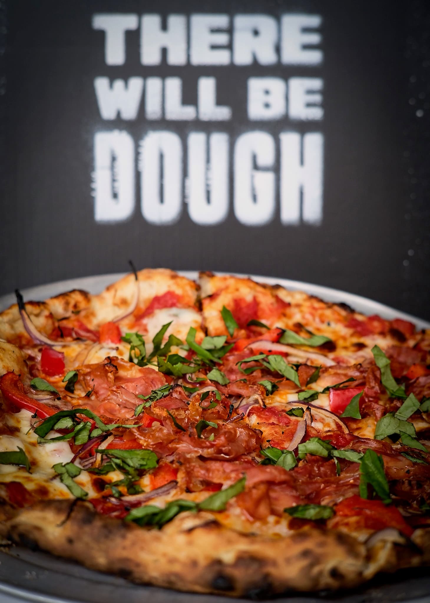 Prosciutto, red onion and basil pie in front of the There Will Be Dough banner
